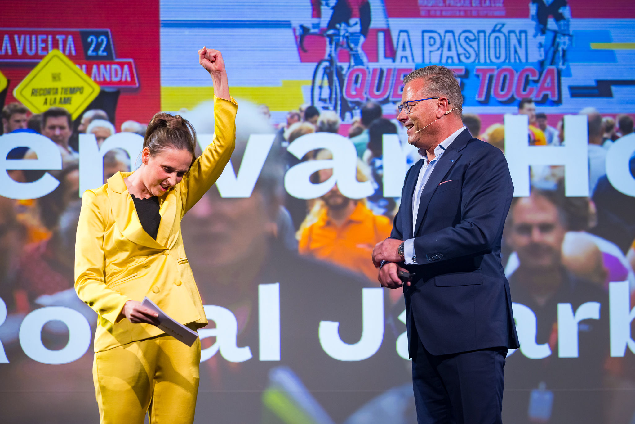 Vrouw gekleed in geel pak en een man gekleed in een blauw pak op een podium.